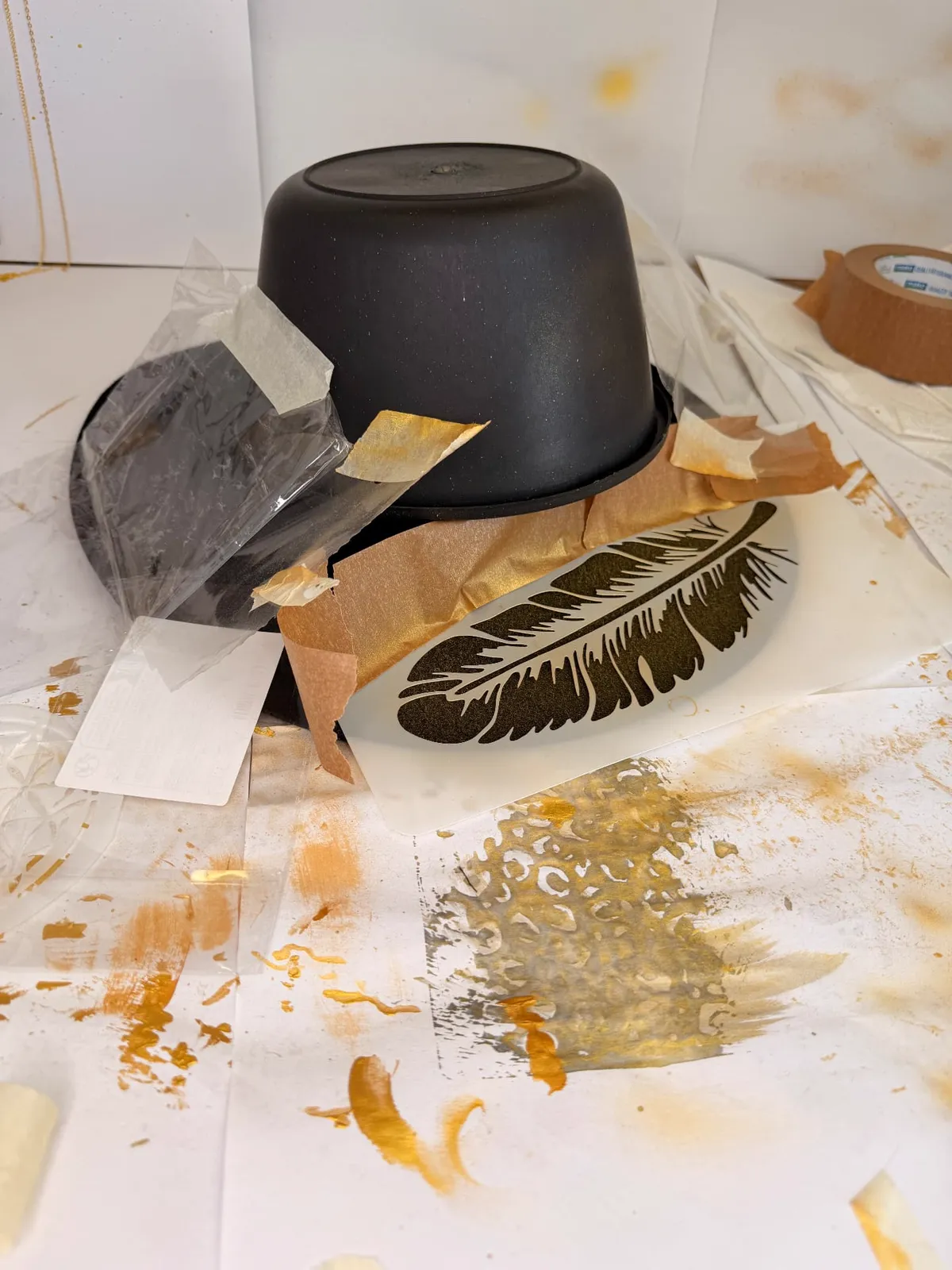 Blank black felt hat masked with tape beside a feather stencil and gold pigment in the studio