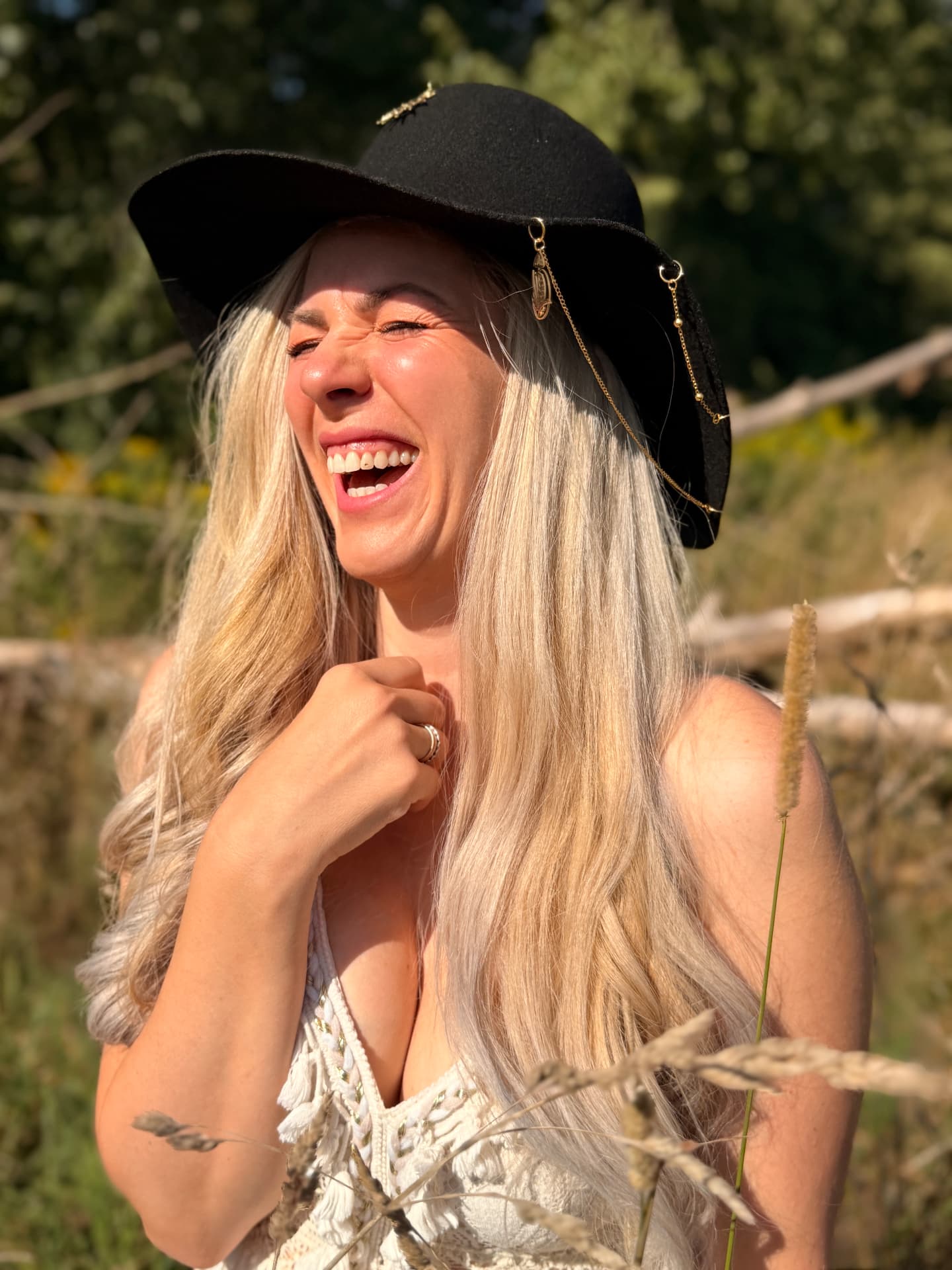WILD & GOLD founder laughing in a sunlit field, wearing a black handcrafted felt hat with gold chain details